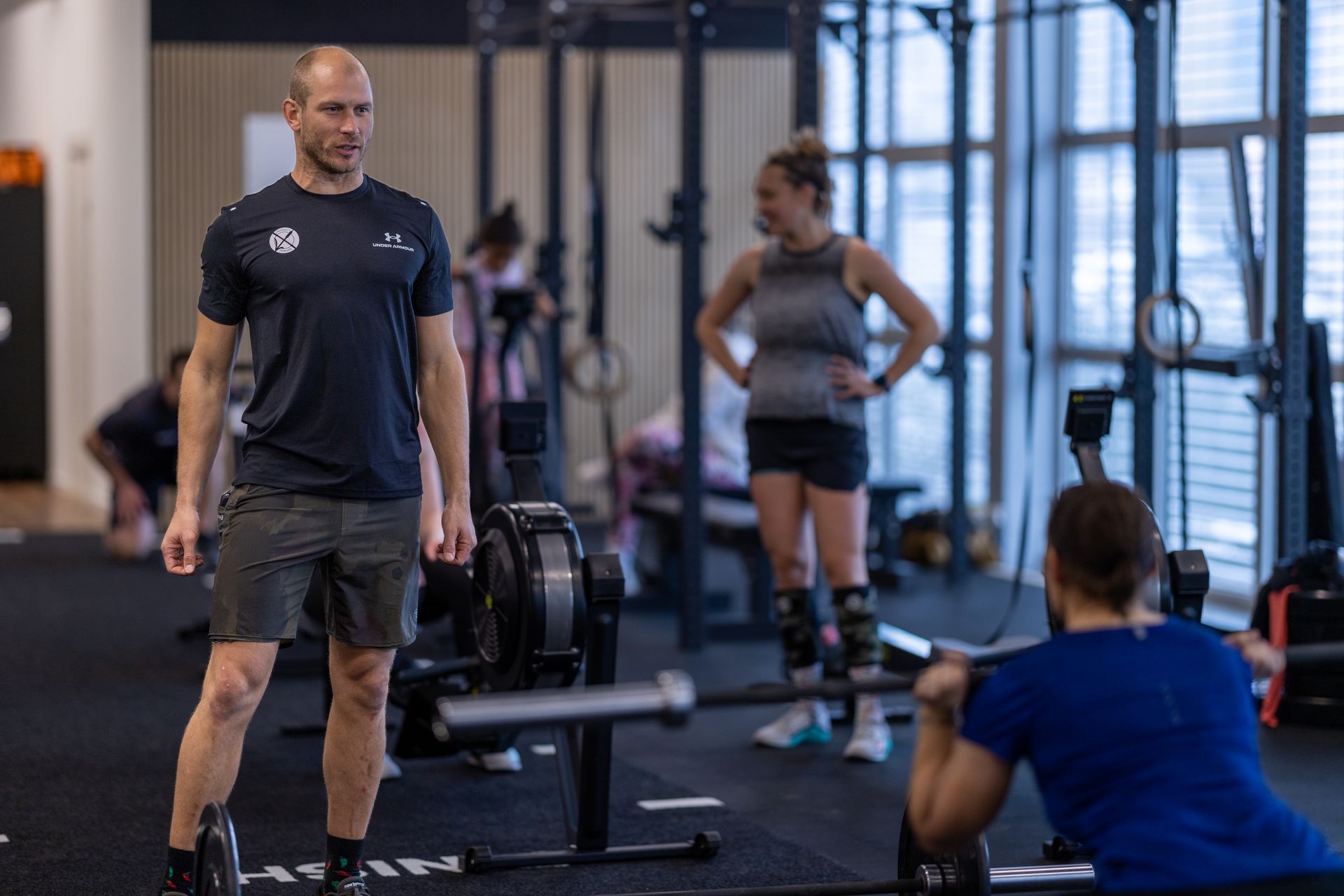 A coach stands in a gym in Winterthur, watching someone squat with a barbell while another person stands nearby