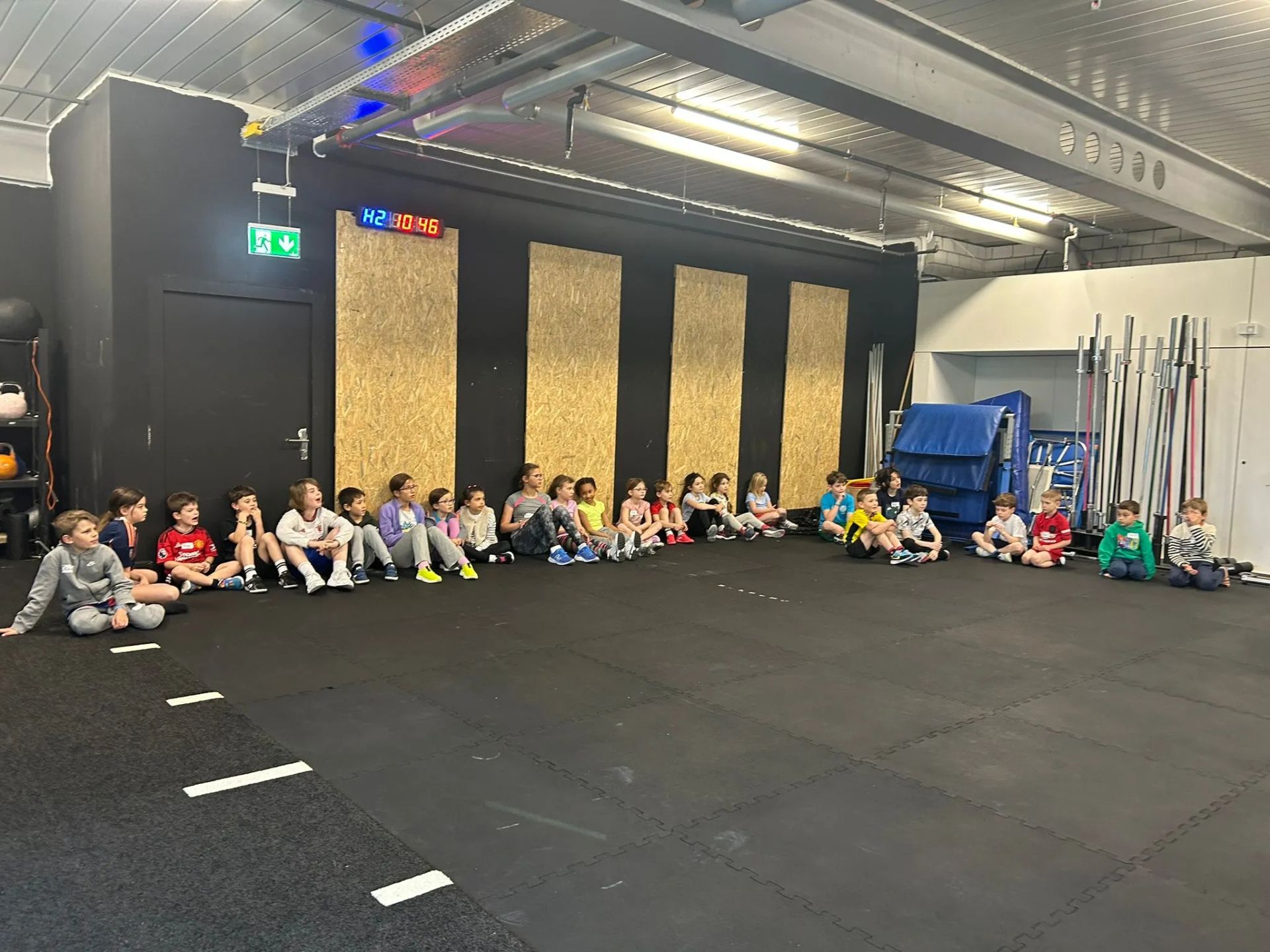 A large group of children sitting on a black gym floor, arranged in a line against a wall with wooden panels.