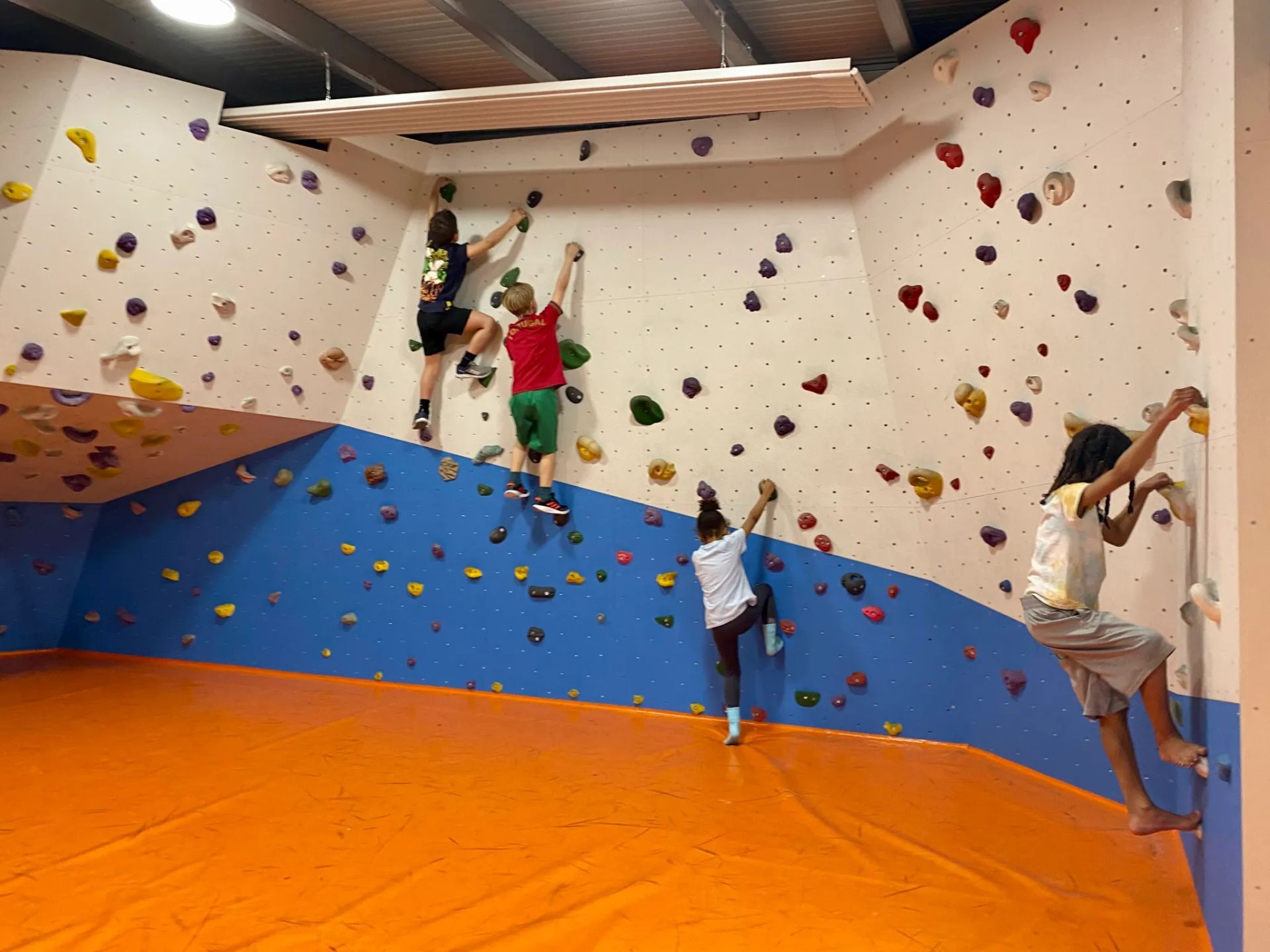 Four people climb a bouldering wall with colorful holds above a bright orange safety mat in an indoor gym.