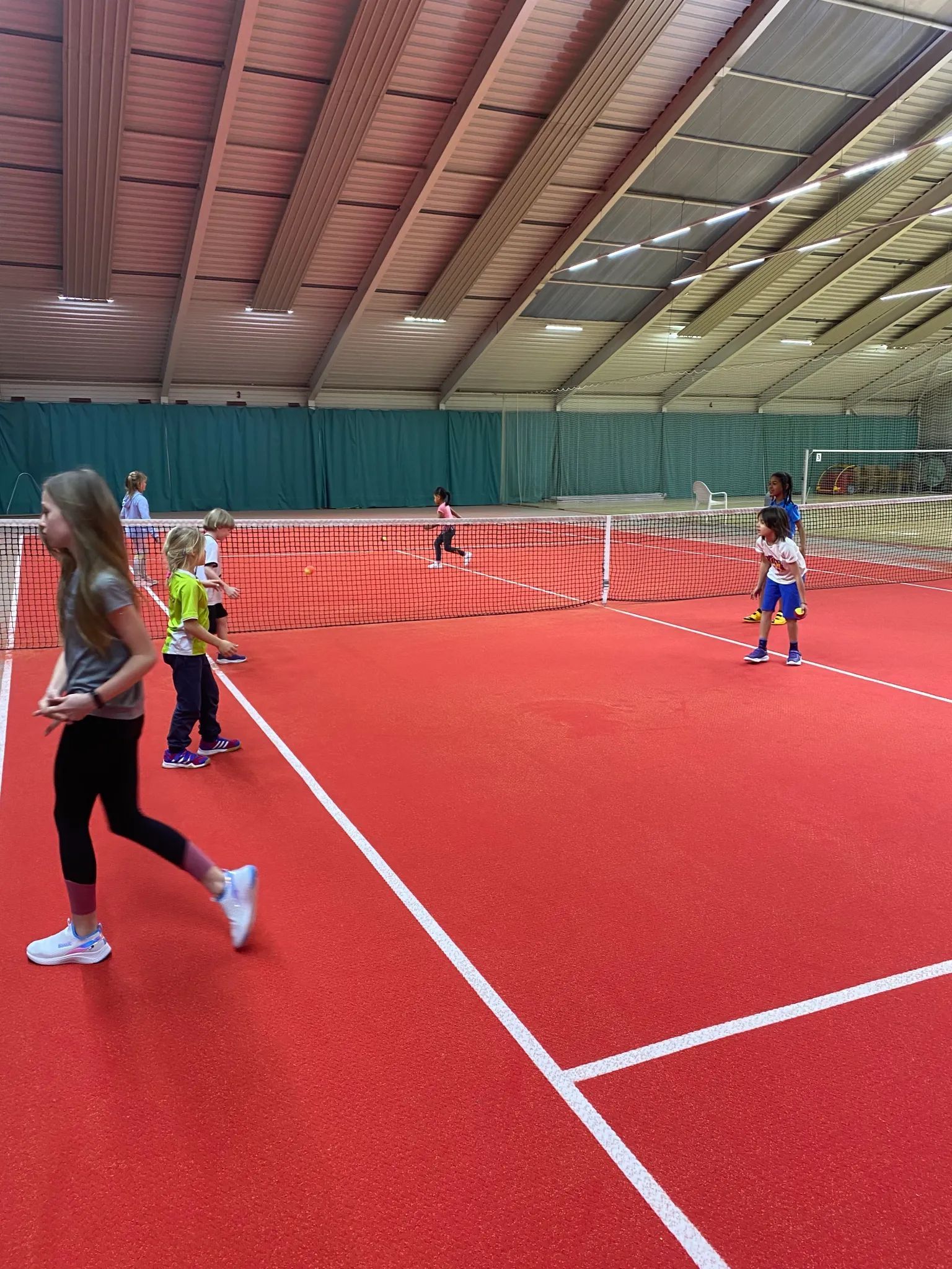A group of children play on an indoor red tennis court under a large metal-framed roof.