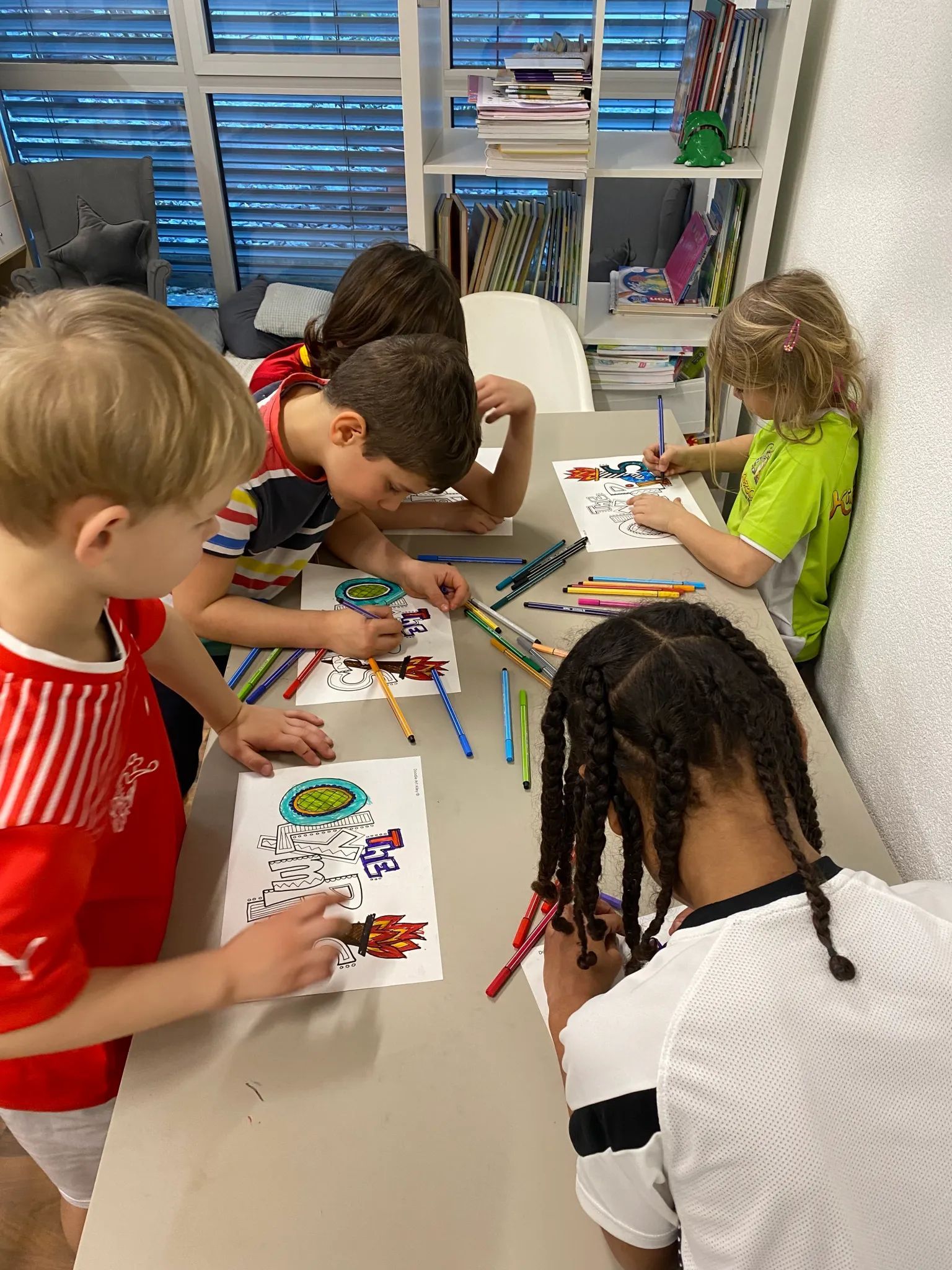 Four children sit at a table in a room, focused on coloring and drawing with markers on paper.