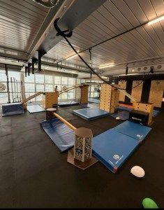 An indoor parkour or gymnastics training facility with wooden platforms, blue foam landing mats, and a suspended rope.