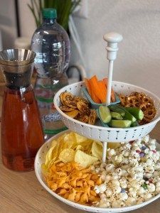 A two-tiered serving tray with snacks, including popcorn, chips, pretzels, and fresh vegetables, next to drinks.