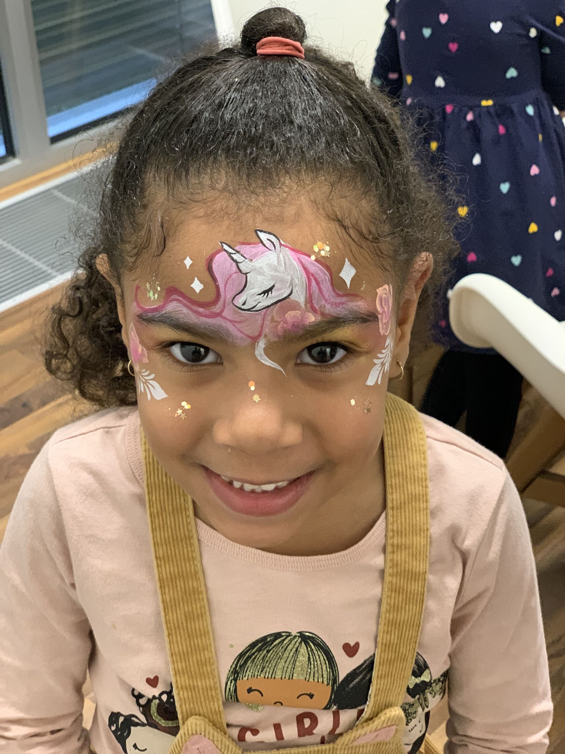 A child smiling with unicorn face paint on their forehead, featuring a white unicorn and pink hair design.