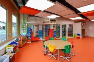 Brightly colored kindergarten classroom with a round table, chairs, storage shelves, and orange flooring.