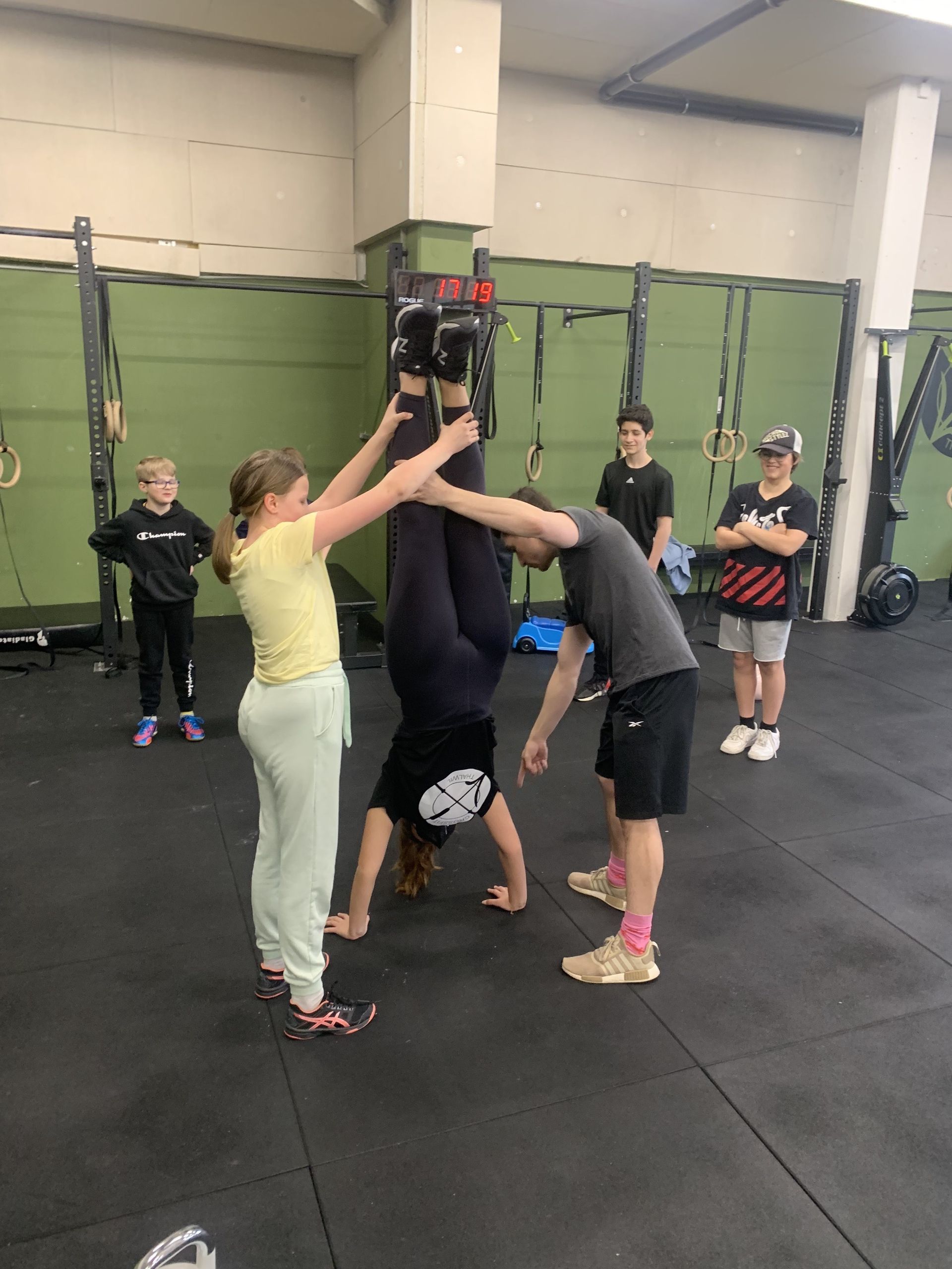 A person in a gym in Winterthur performs a handstand while two people assist by holding their legs upright.