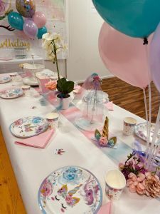 A party table set for a unicorn-themed event, with themed plates, napkins, cups, a unicorn centerpiece, and balloons.
