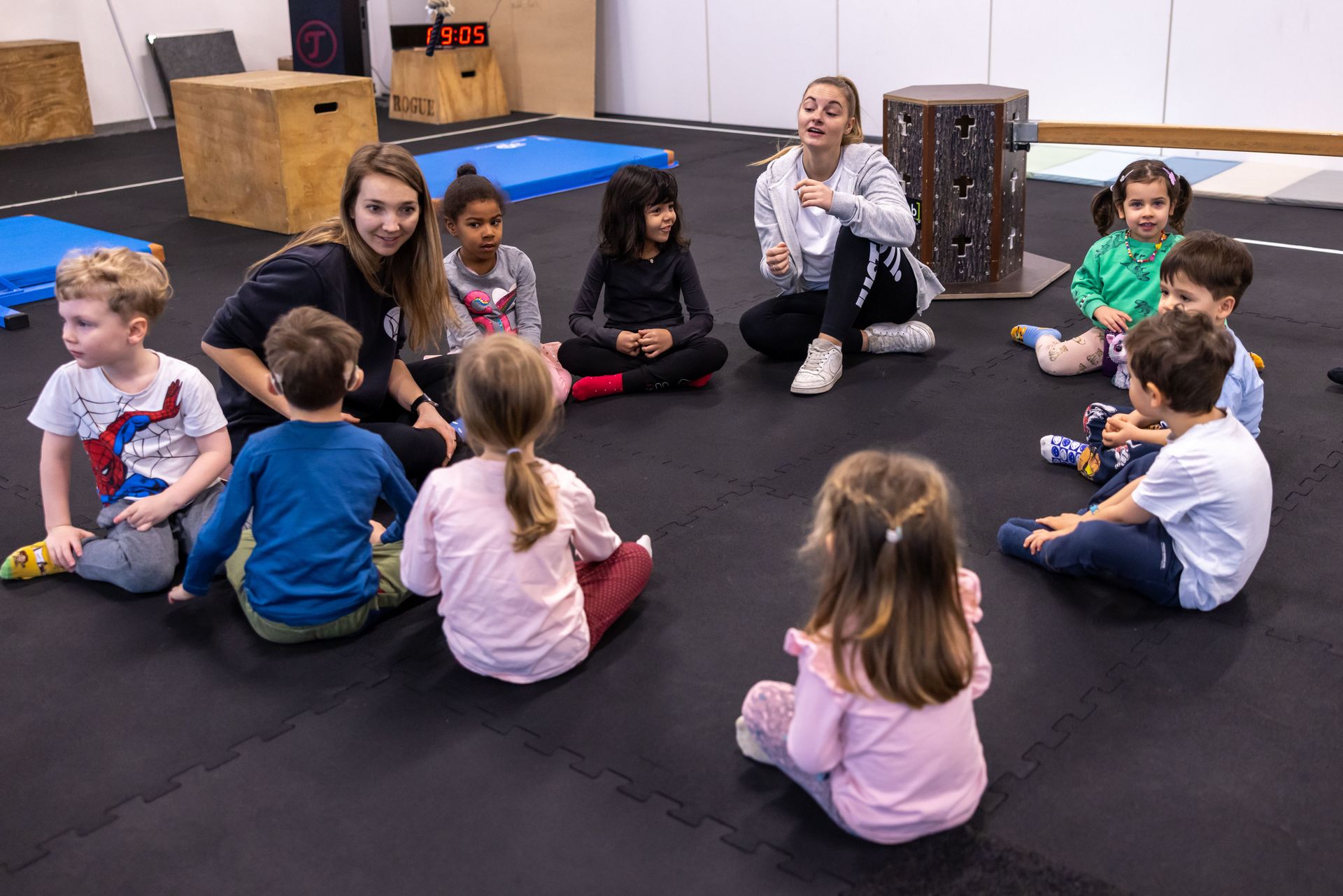 A group of children and two instructors sitting in a circle on a gym mat during a class.