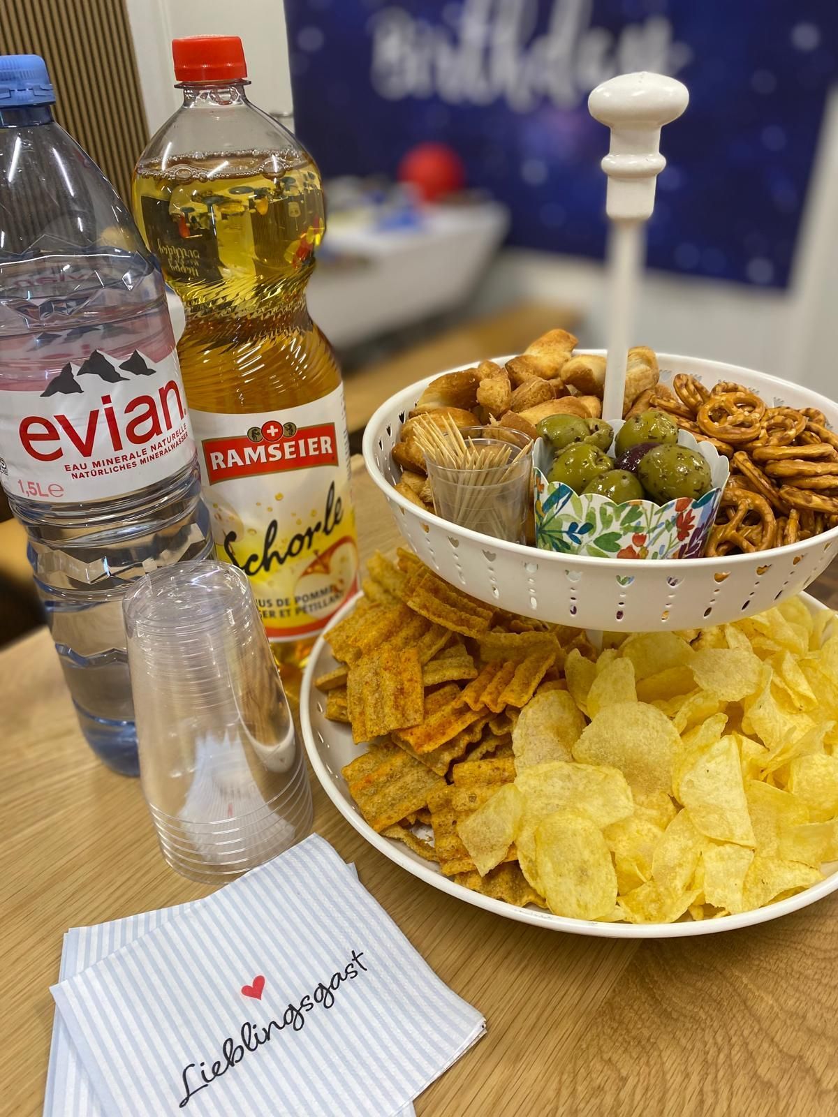 A tiered serving tray holds snacks, olives, and pretzels, next to bottles of Evian and Schorle on a wooden table.