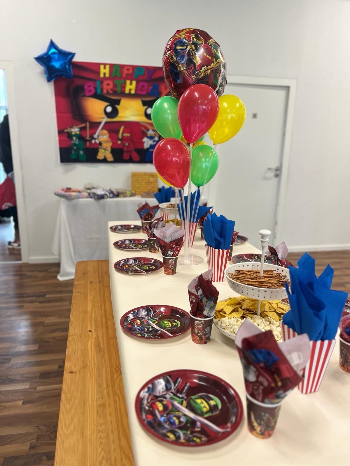 A party table set with Ninjago-themed plates and cups, a balloon bouquet, and snacks in front of a birthday backdrop.