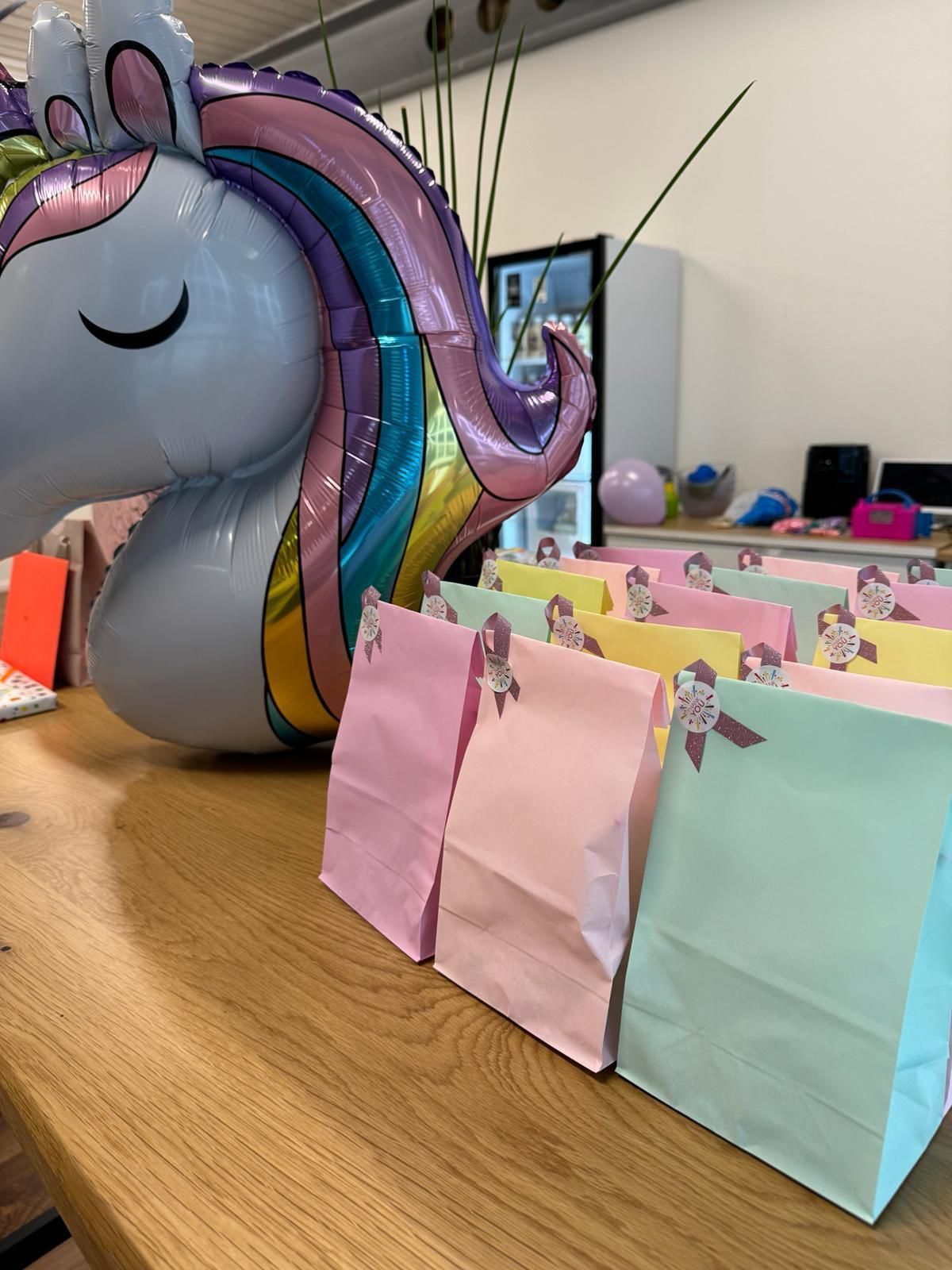 A row of pastel-colored gift bags arranged on a wooden table next to a large, colorful unicorn-shaped foil balloon.