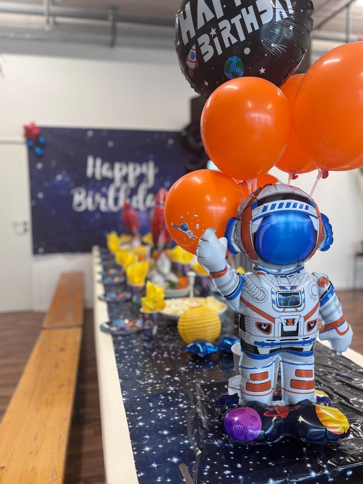 An astronaut balloon centerpiece and orange balloons sit on a long, space-themed table with a Happy Birthday backdrop.