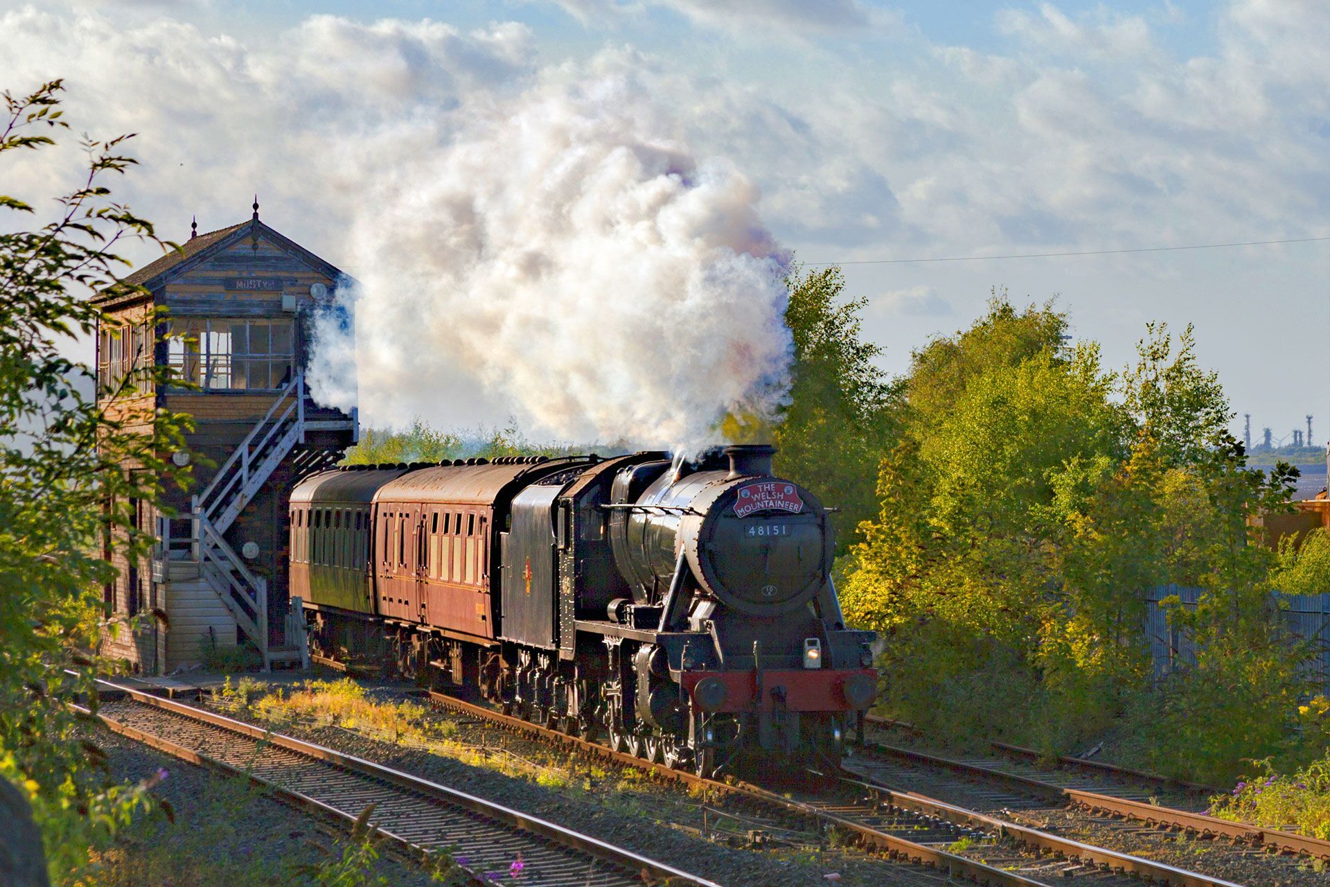 Steam locomotive pulling vintage cars past a signal box, with white smoke billowing through a sunny green landscape.