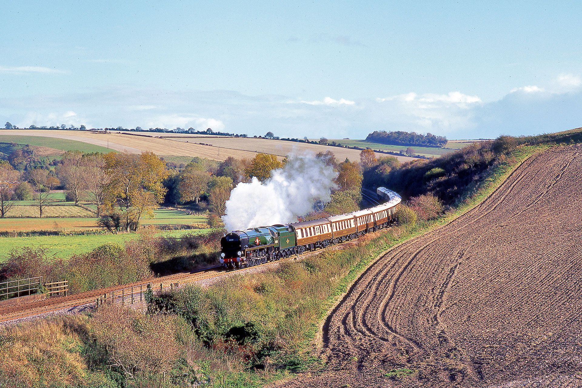 Steam train curving through a rural hillside, white smoke rising over green fields and brown earth