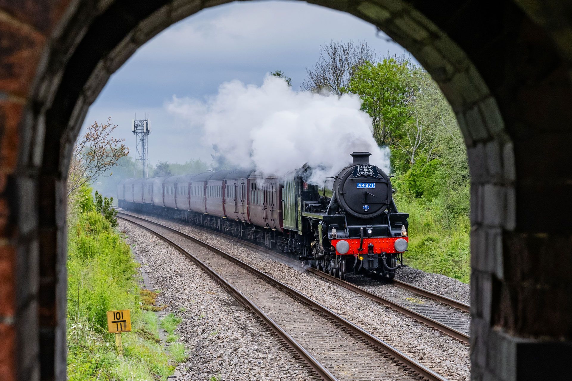 Steam locomotive pulling carriages through an arched tunnel toward the viewer, billowing white smoke.