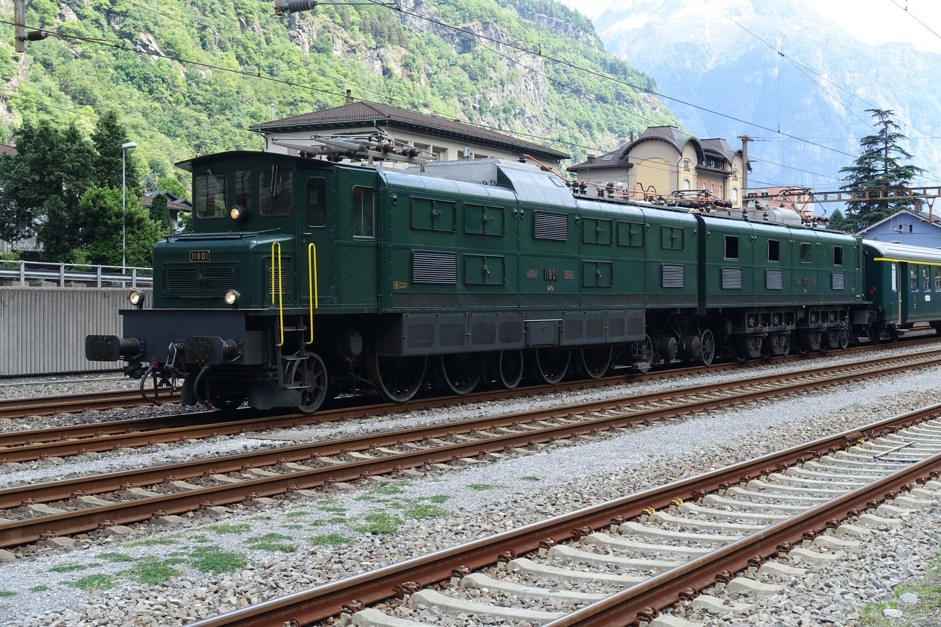 Dark green vintage train on tracks at a station with mountains and trees in the background