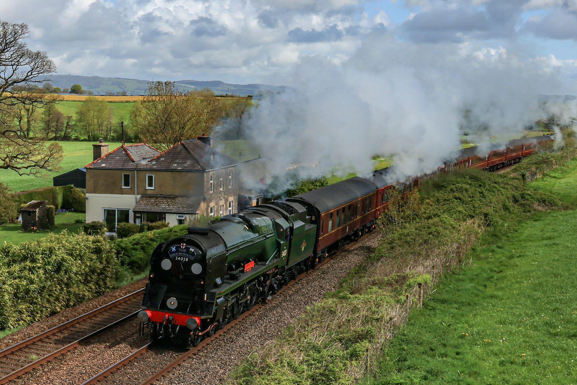 Steam locomotive pulling passenger cars through green countryside beside a rural house