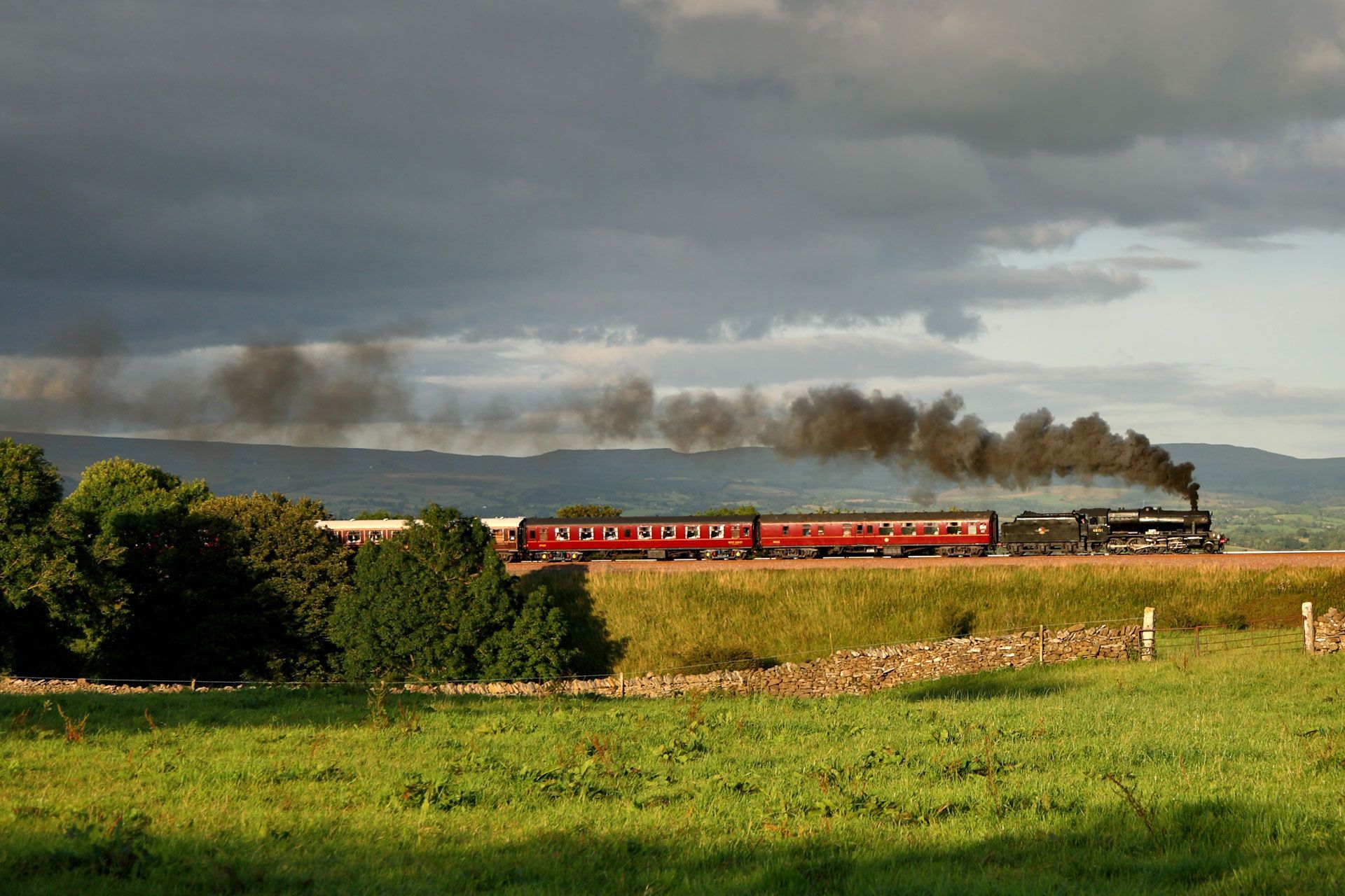 Red passenger train crossing a green countryside under dark storm clouds