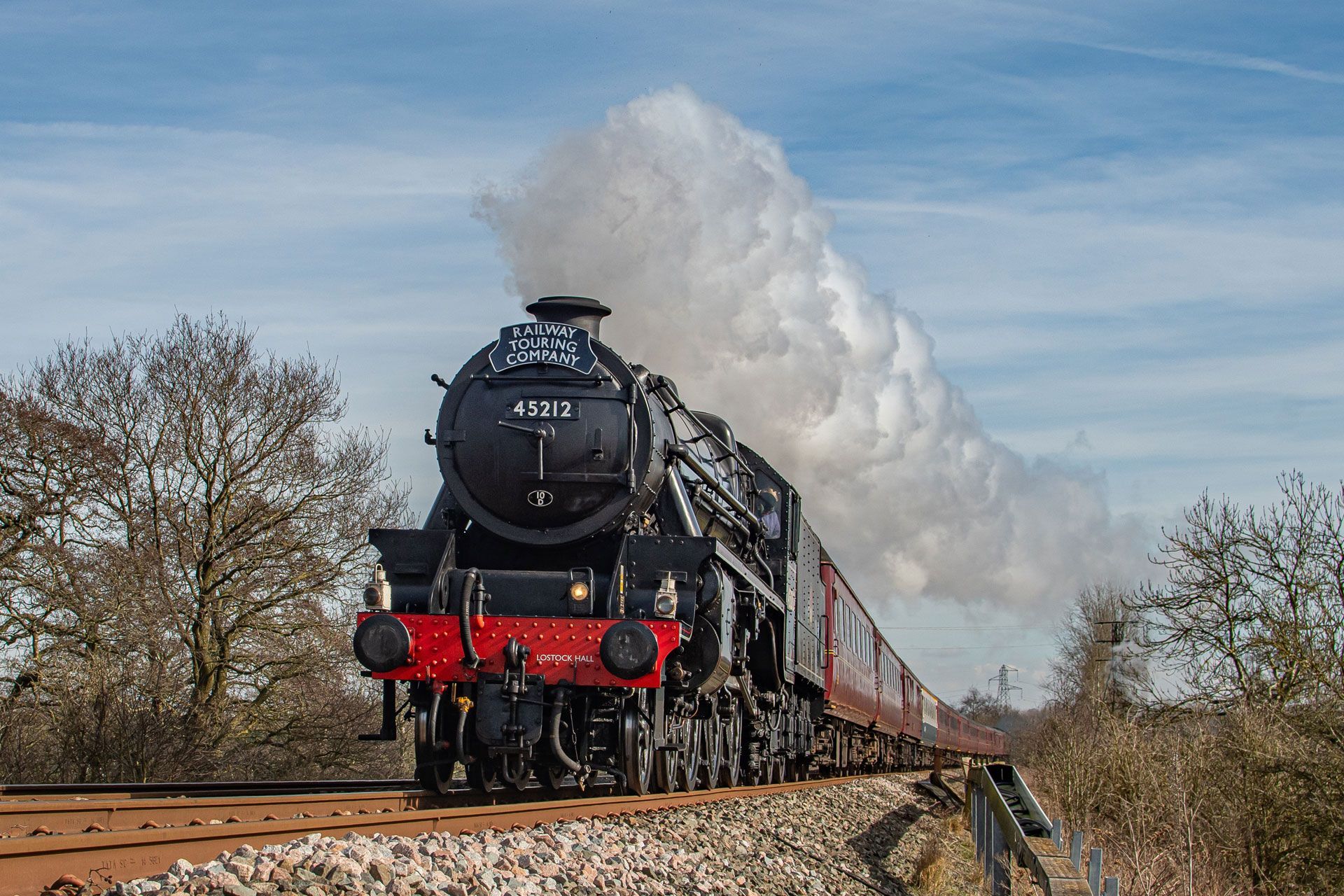 Black steam locomotive pulling passenger cars through a rural landscape, with billowing white smoke.