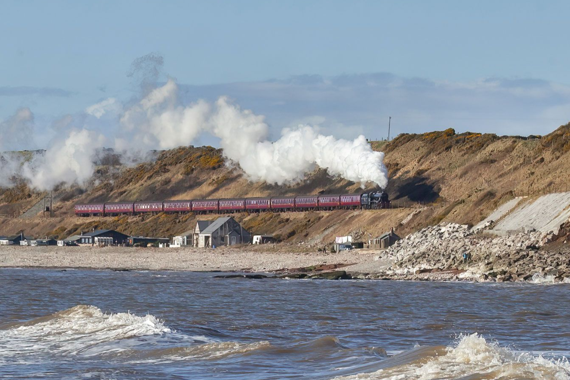 Red train crossing rocky seaside cliffs with steam rising above the shore