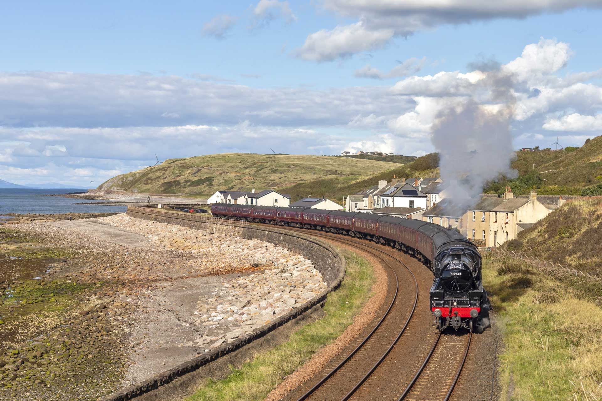 A steam train pulling dark passenger cars travels along a coastal track near a small village under a cloudy blue sky.