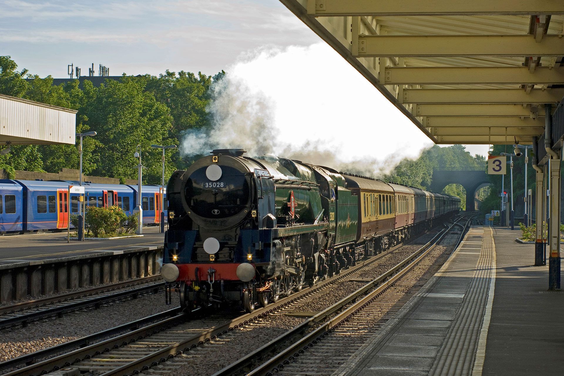 Steam locomotive pulling passenger cars into a station under a platform canopy, with steam rising.