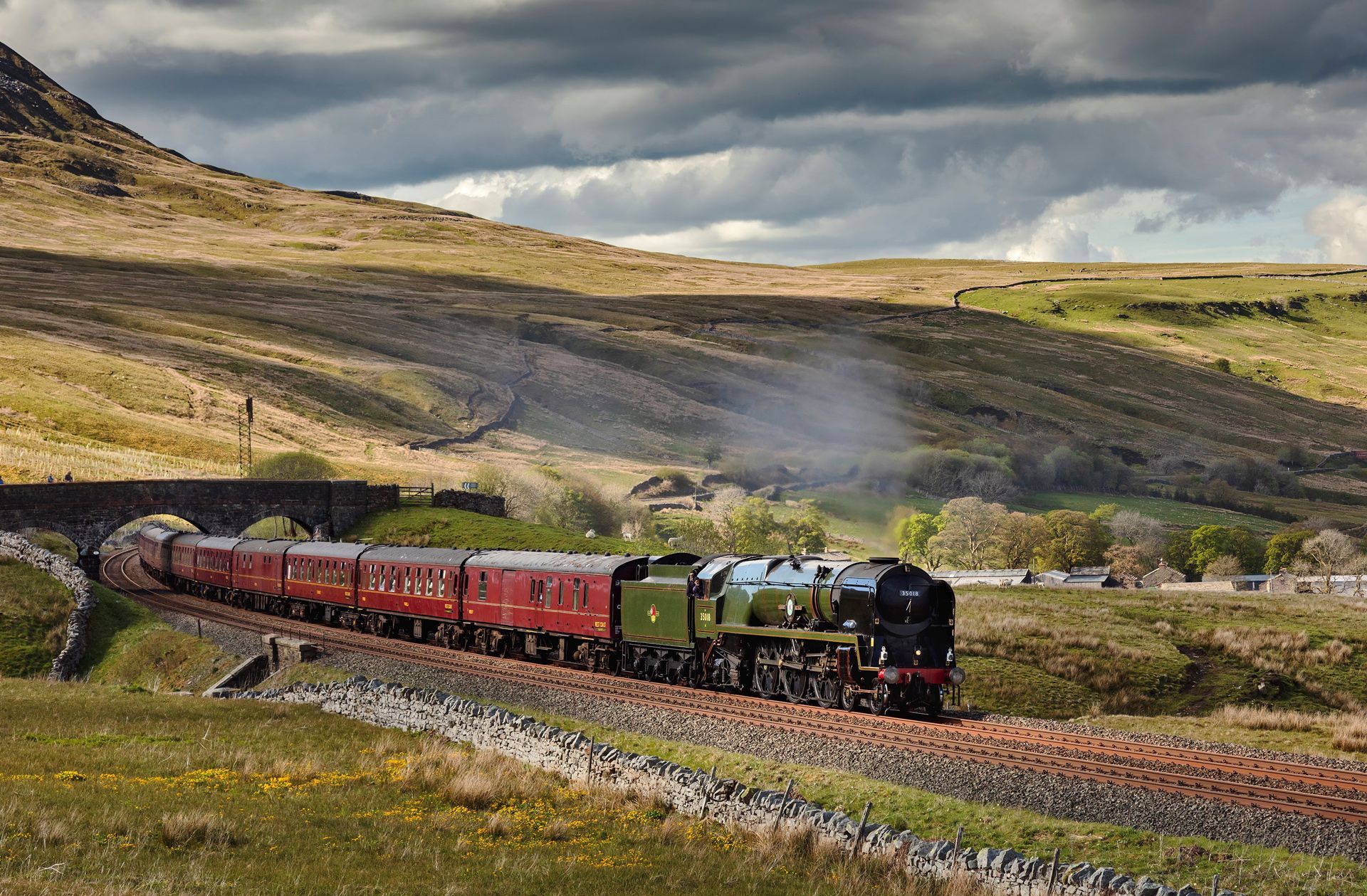 Steam locomotive pulling red passenger cars through a grassy hillside under cloudy skies