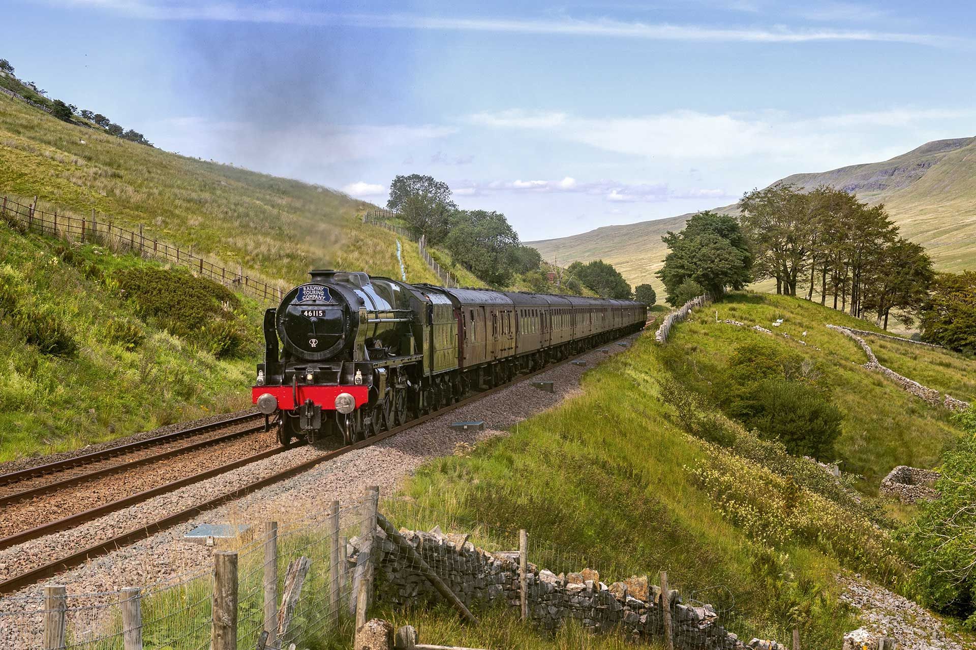A steam train travels through a lush green valley with rolling hills on a bright, clear day.