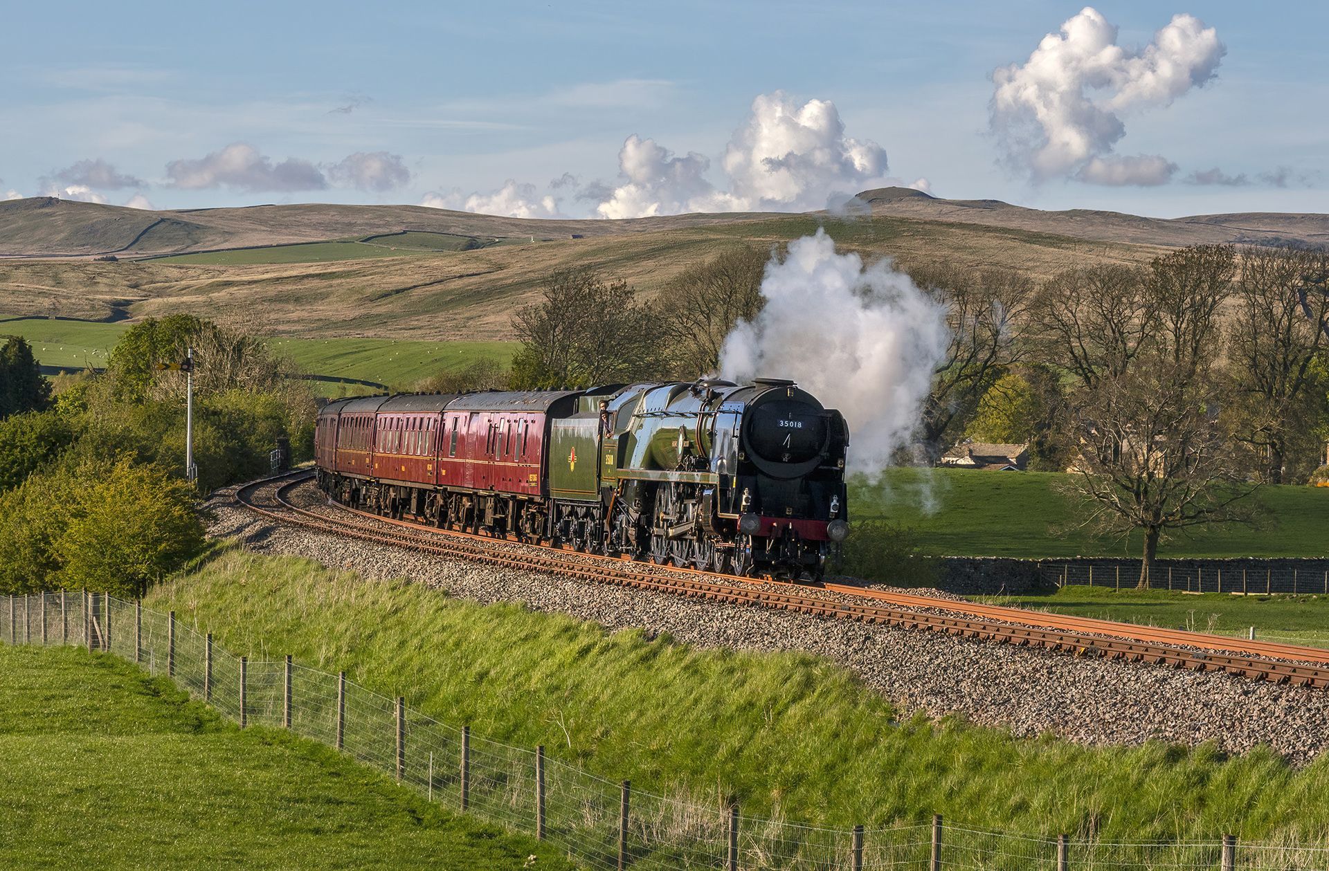 Steam locomotive pulling red coaches through a rural valley, with white steam against hills and clouds.