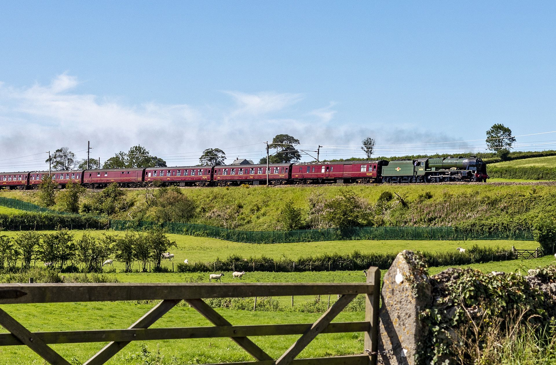 Red train crossing green countryside under blue sky, with a wooden gate in the foreground