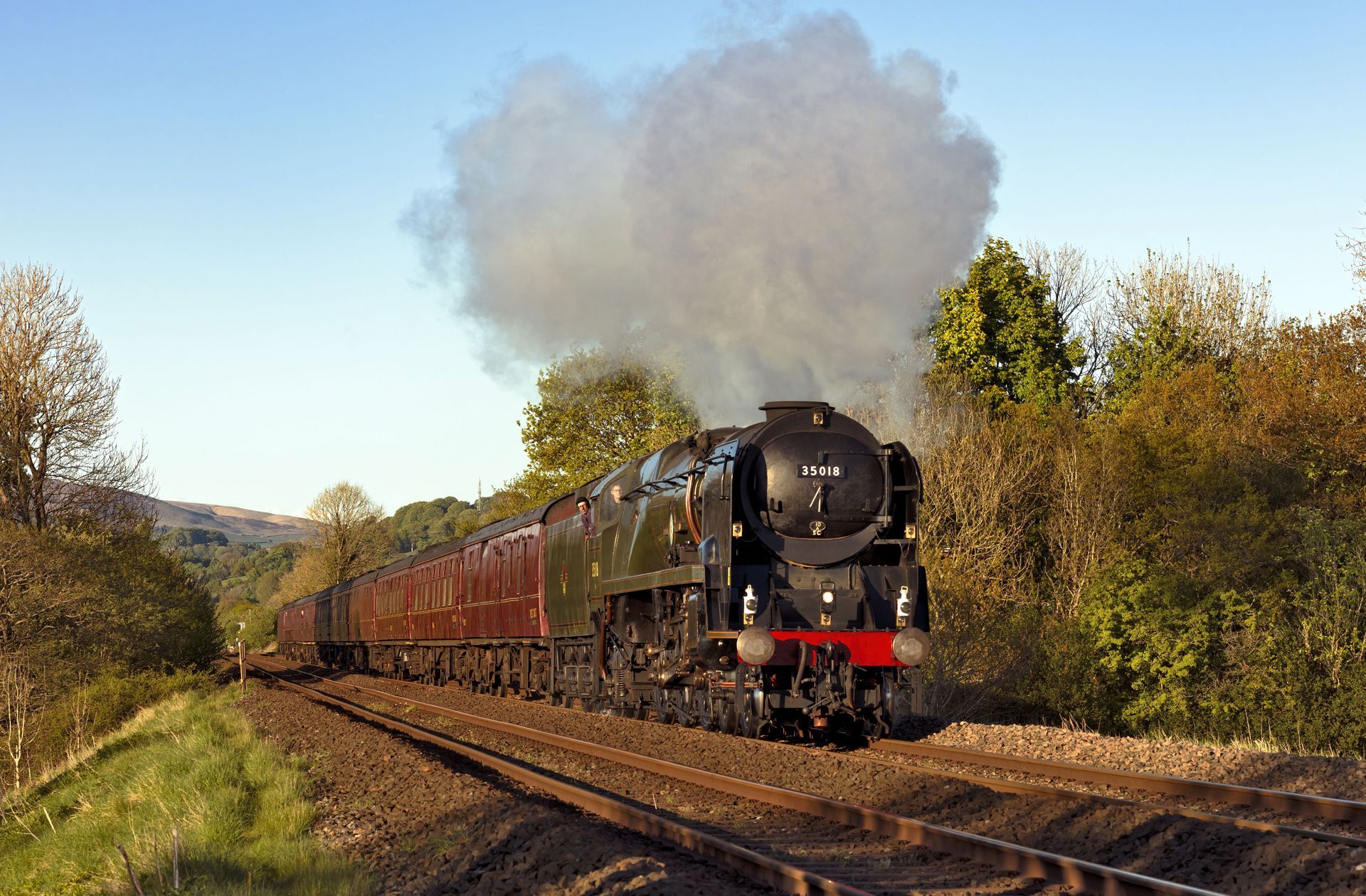 Black steam locomotive pulling red passenger cars along a rural track under a plume of smoke.