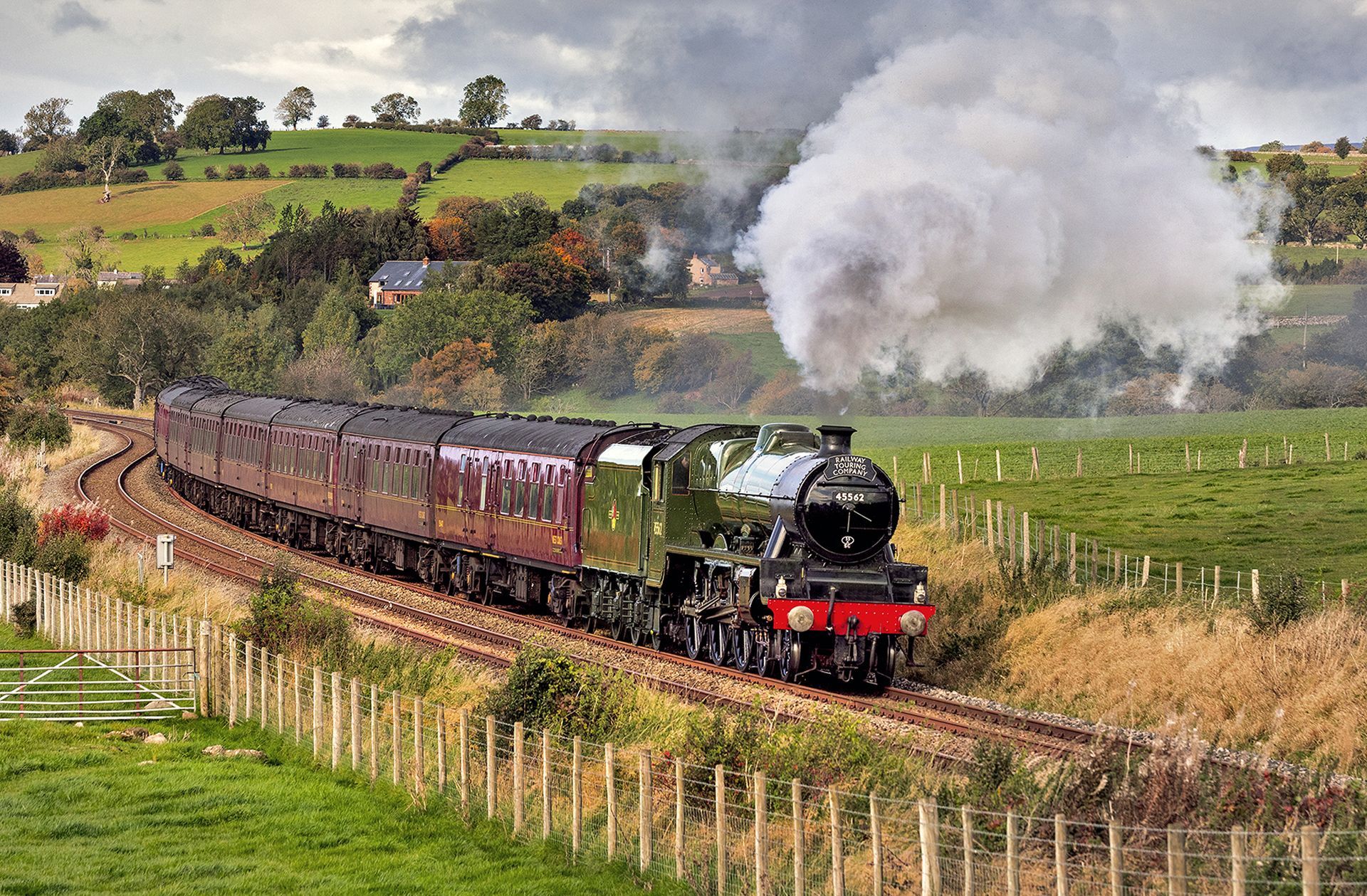 Steam locomotive pulling maroon coaches through a green countryside, with white smoke billowing overhead