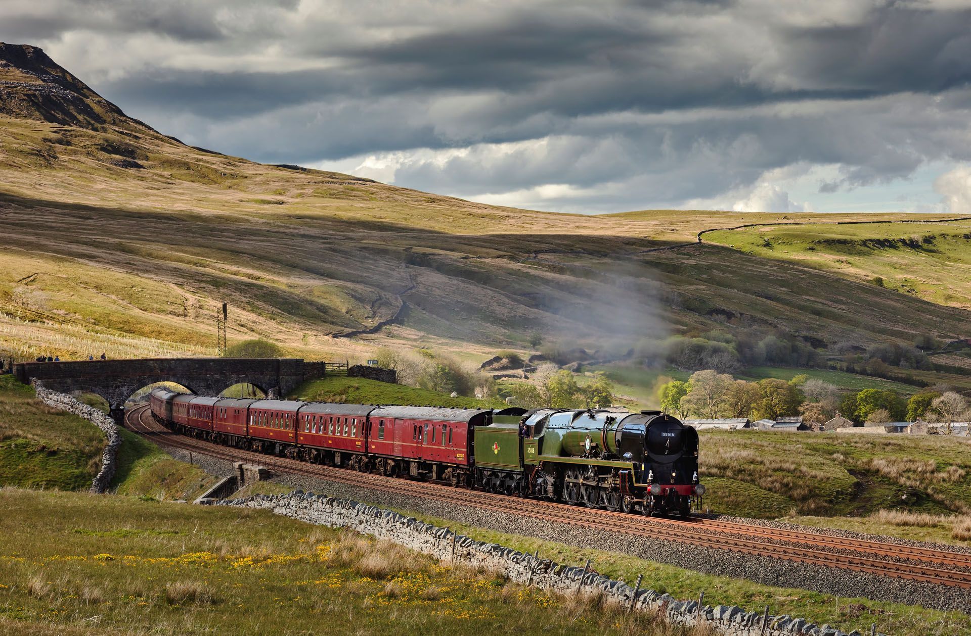 Steam train with red carriages crossing a grassy hillside under cloudy skies