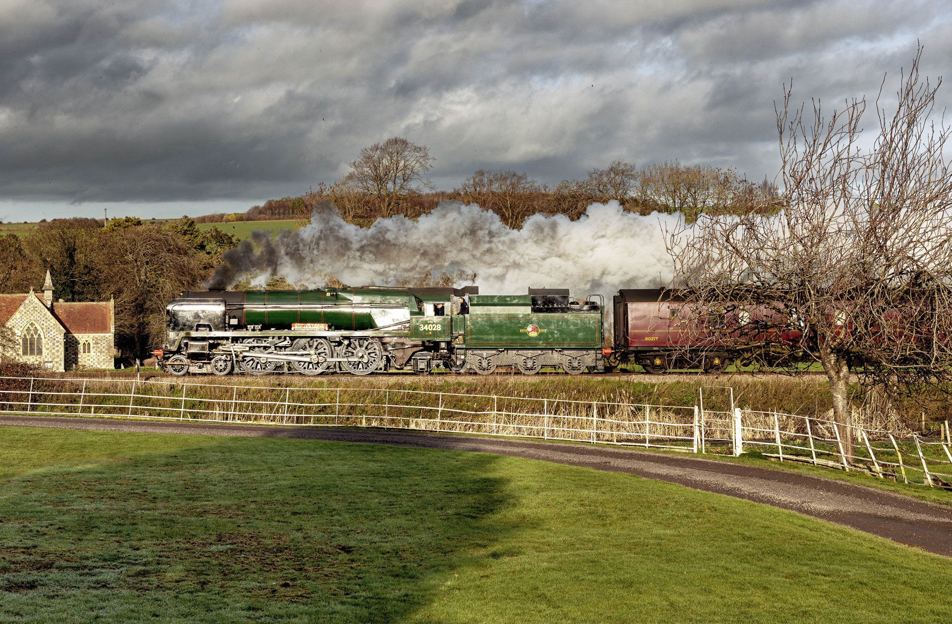Steam train crossing a countryside viaduct under cloudy skies, with green fields in the foreground