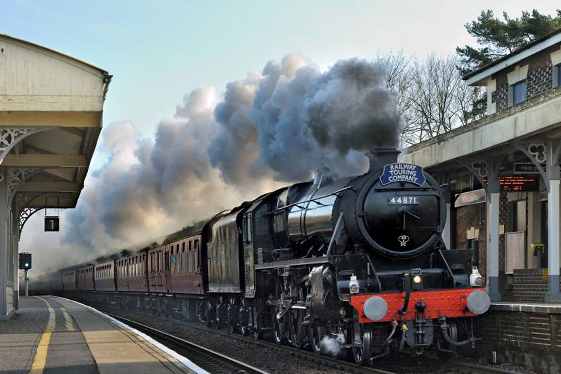 Black steam locomotive pulling a train through a station, billowing smoke under an overcast sky