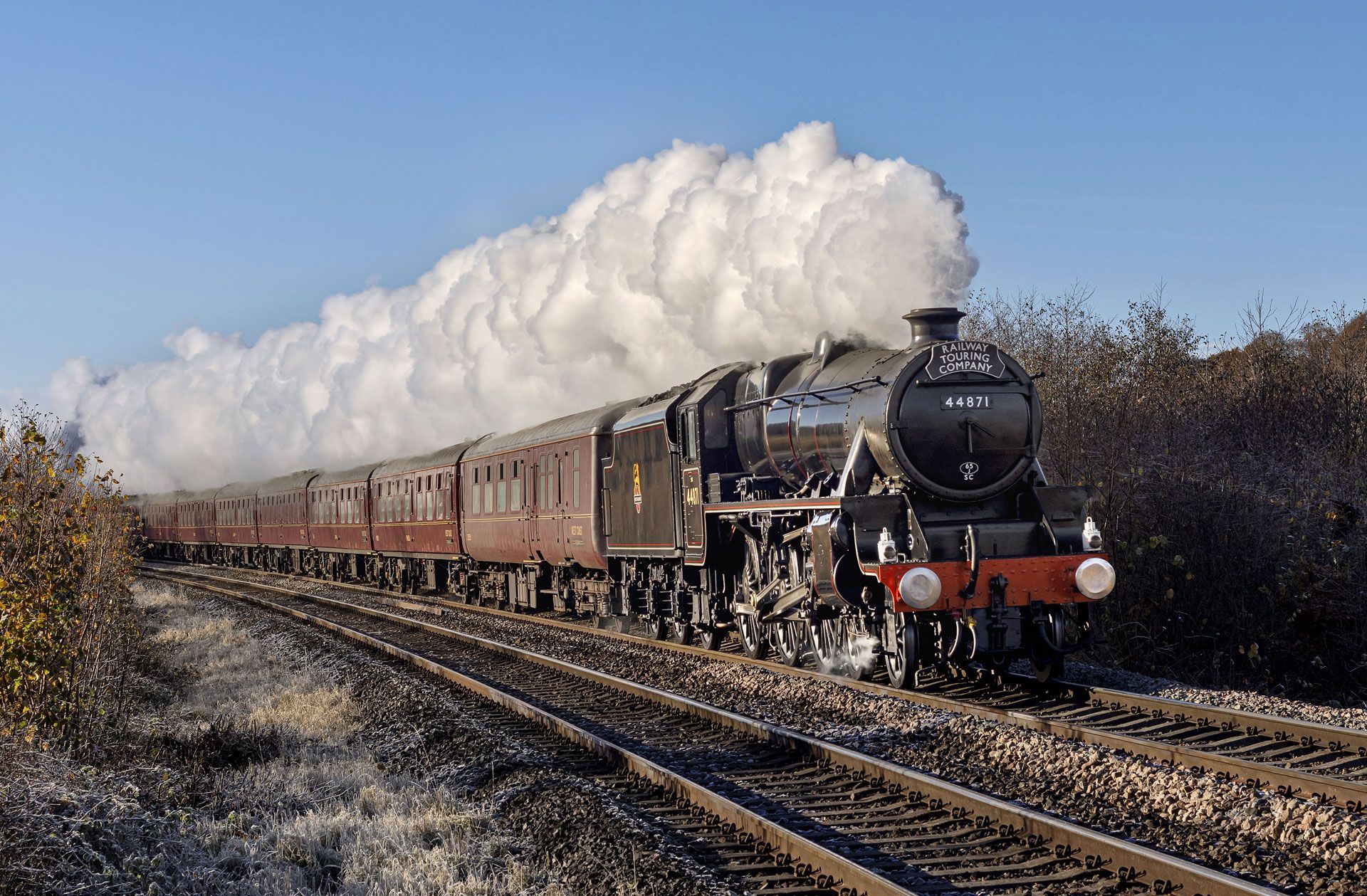 Steam locomotive pulling a long passenger train through a rural landscape, leaving a plume of white smoke