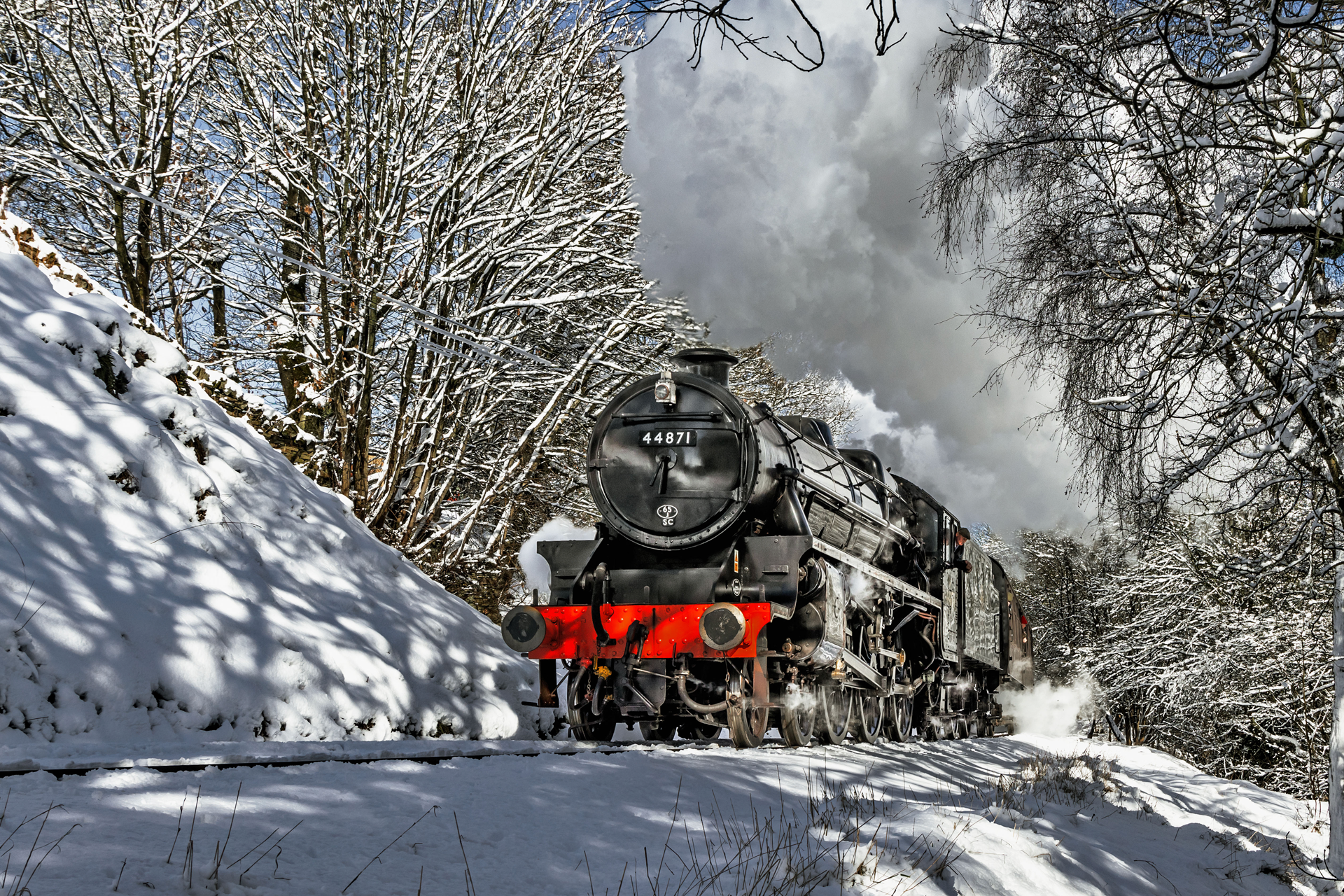 Steam locomotive driving through a snowy forest with red front plates and smoke billowing above