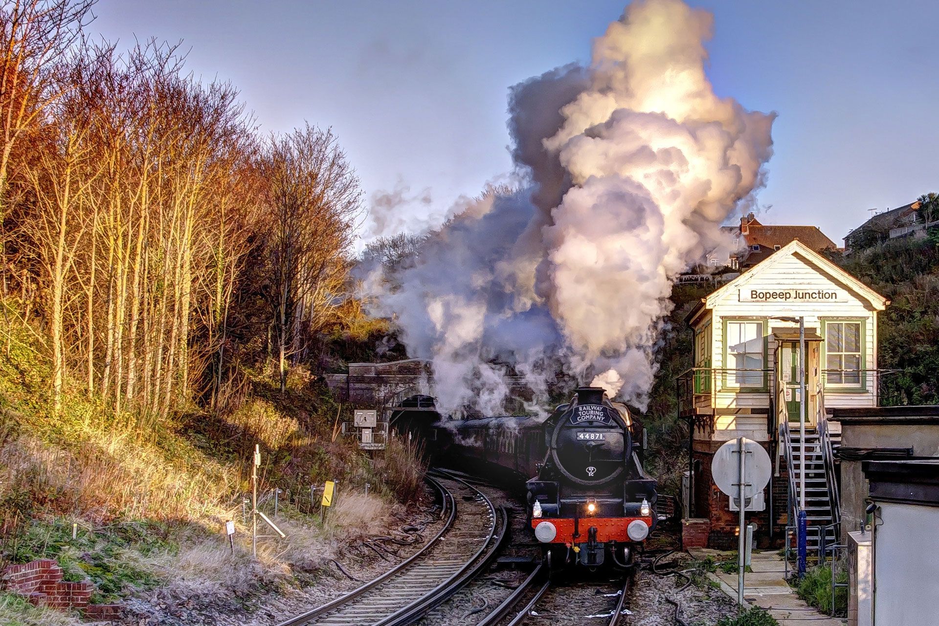 Steam locomotive chugs through a wooded curve beside a small station, billowing white smoke at sunset