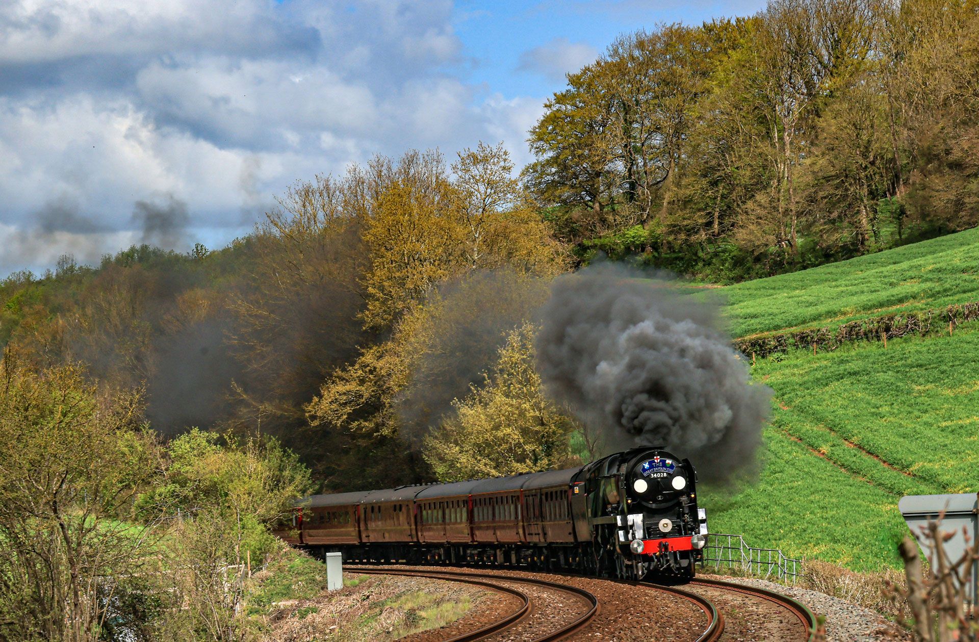 Steam locomotive curving through a green countryside, pulling brown coaches under a cloudy sky
