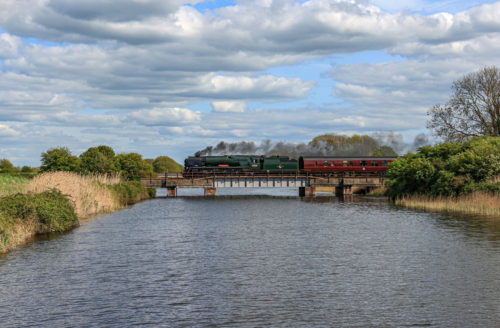 Steam train crossing a bridge over a river under a cloudy sky