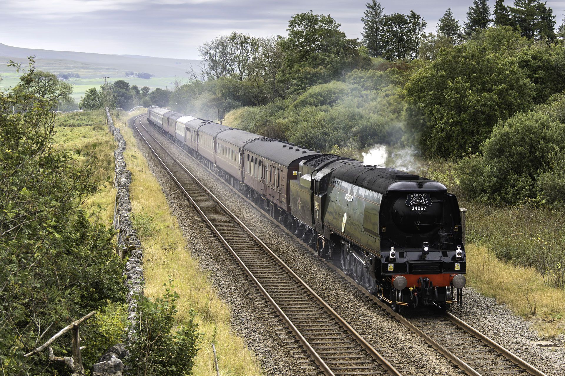 Black steam train pulling passenger cars through green countryside on double tracks