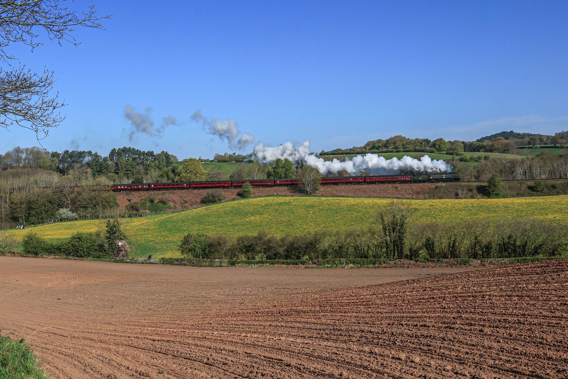 Red steam train crossing a rural field under a blue sky