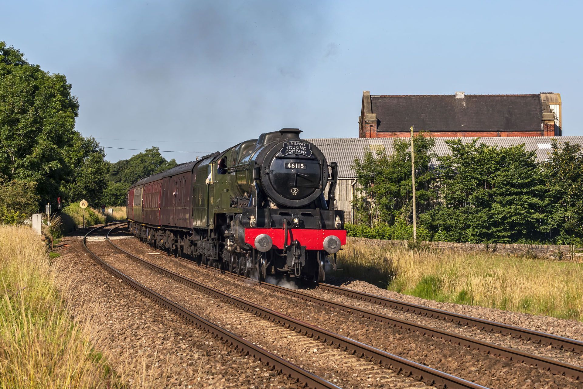 Black steam locomotive pulling maroon coaches along rural tracks beside trees and a house