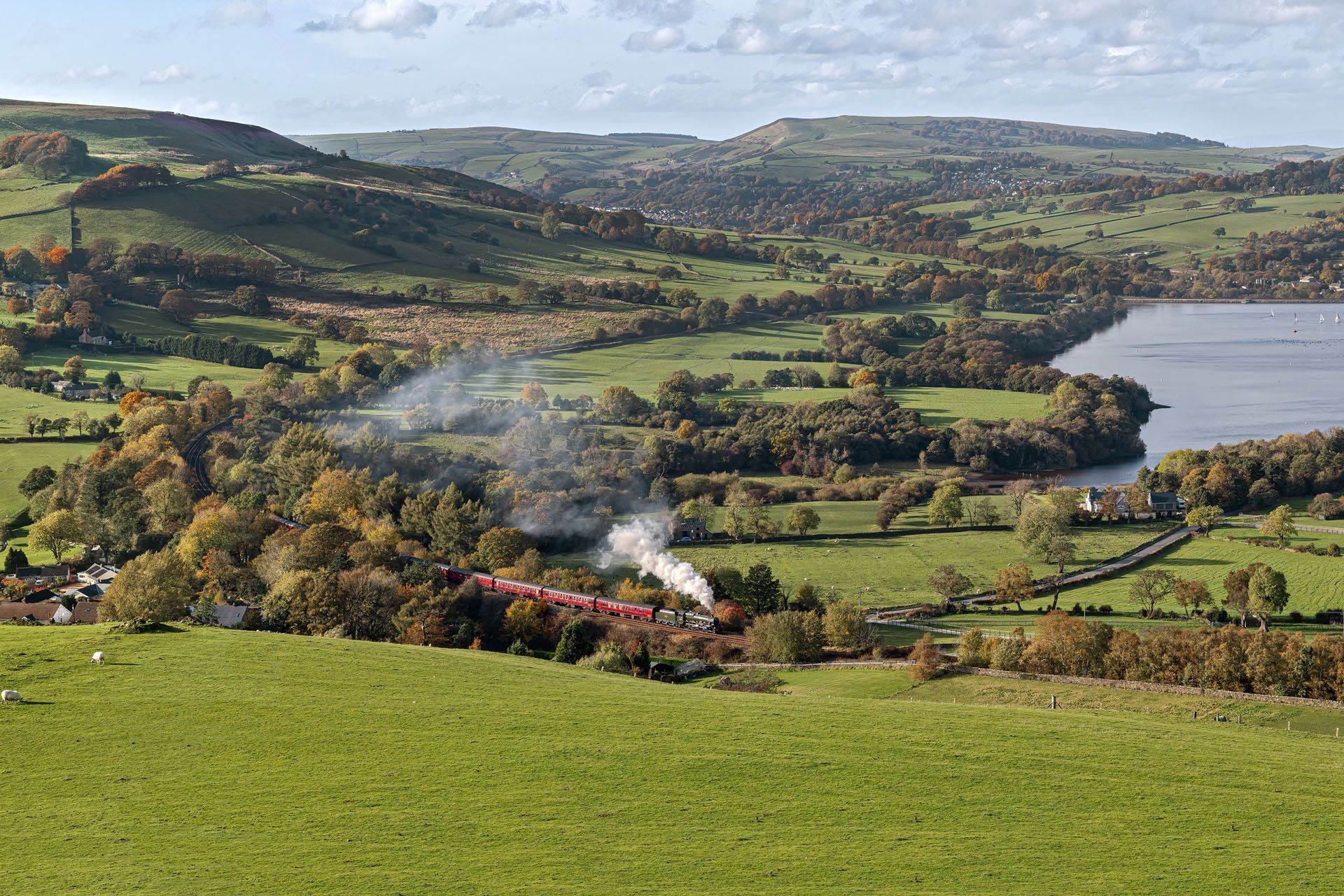 Green valley landscape with hills, fields, a lake, and a small train releasing smoke