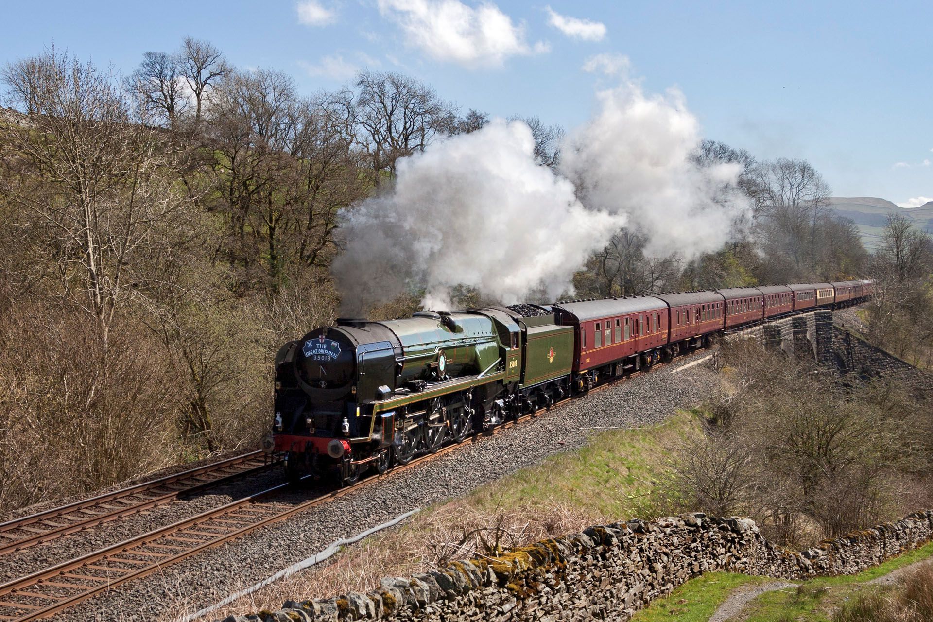 Steam locomotive pulling red passenger cars through a rural landscape with smoke billowing behind