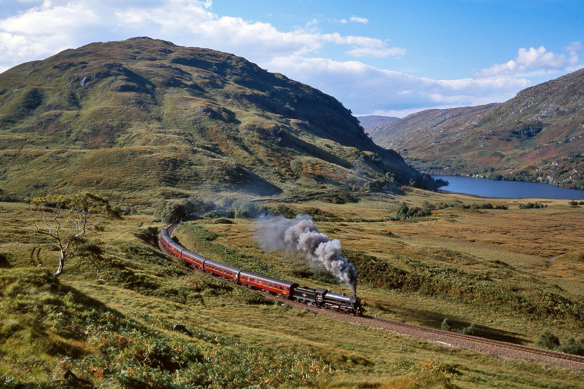 Steam train on a curved track through a grassy mountain valley beside a blue lake