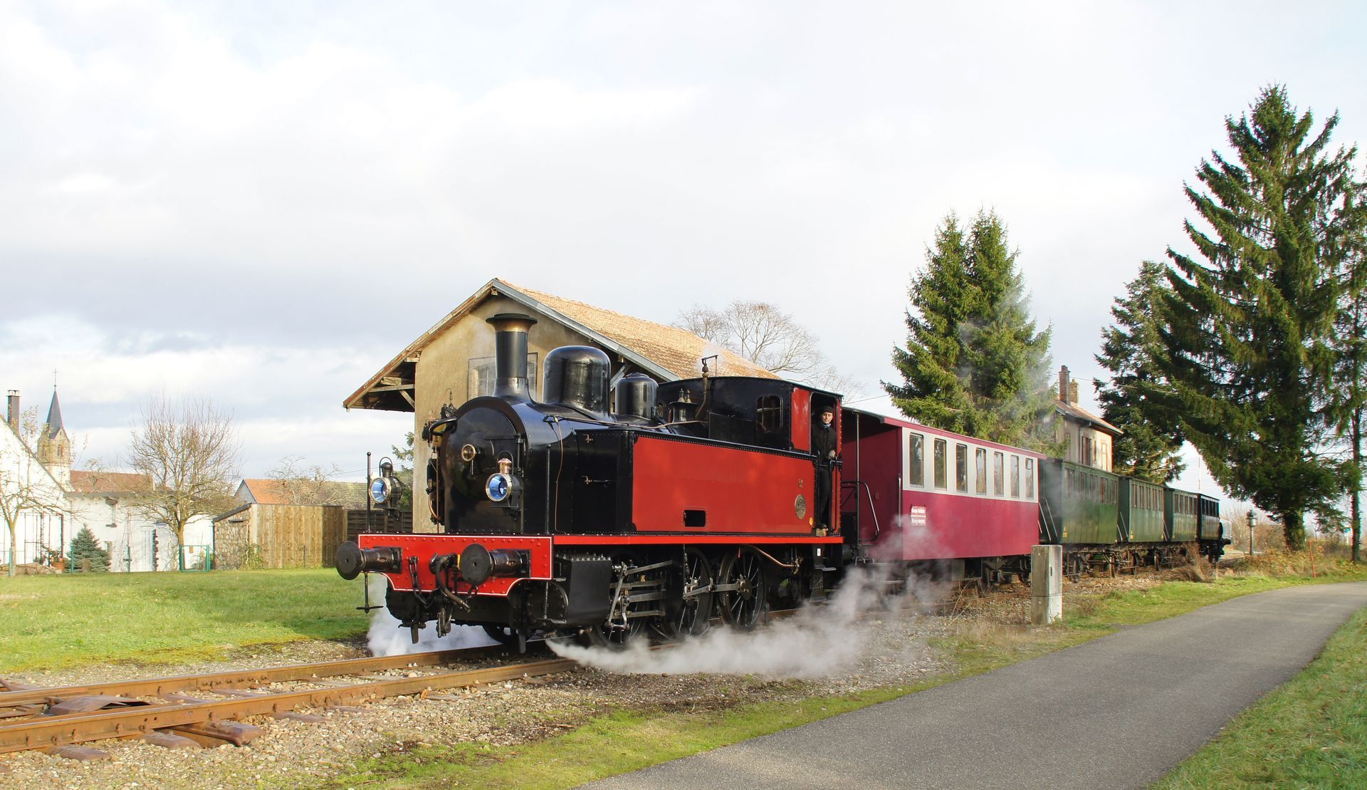 Steam locomotive pulling vintage passenger cars past a small station under cloudy skies