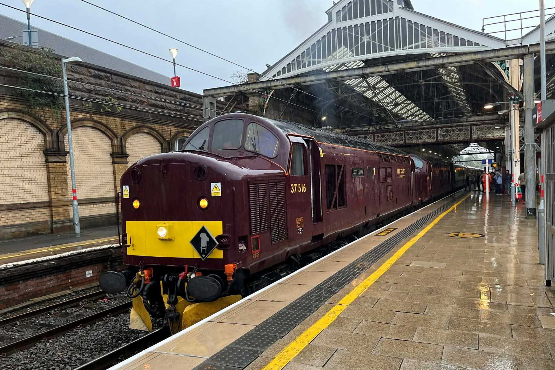 Maroon diesel locomotive at a rainy train station platform under a steel canopy