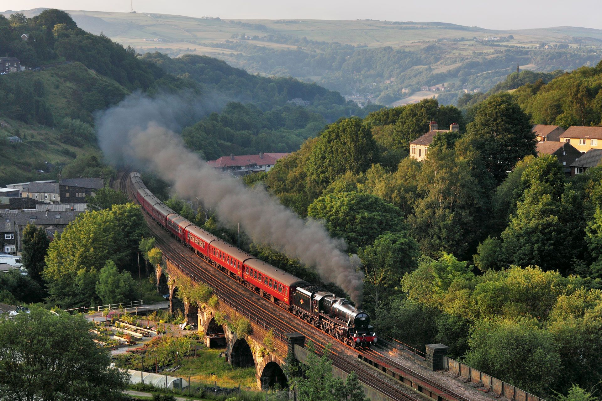 Steam train crossing a stone viaduct through a green valley at sunset