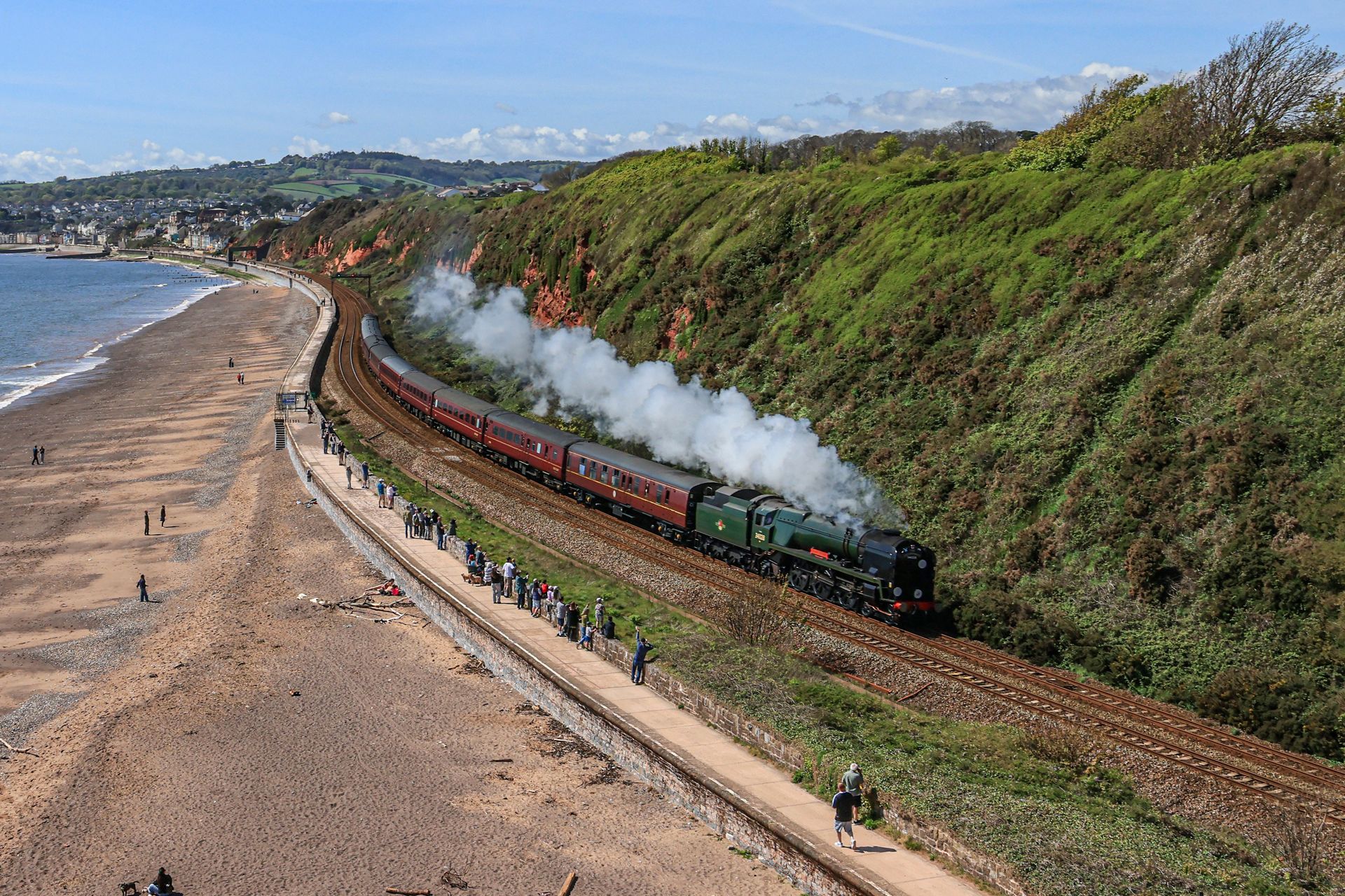 Steam train on a coastal track beside a beach, billowing smoke past green cliffs under a blue sky